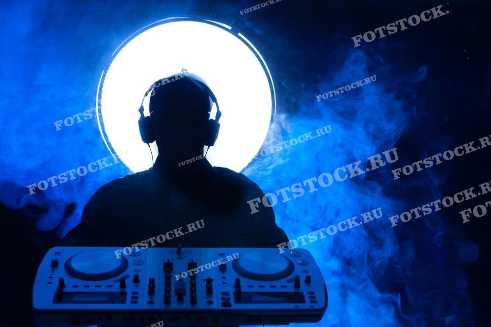 На этой фотографии — силуэт человека в наушниках, стоящего перед DJ-консолью. Вокруг него расстилается дым, окрашенный в синий свет, а за спиной — большой светлый круг, создающий контраст и фокусируя внимание на силуэте. Это изображение отлично подходит для музыкальных мероприятий, рекламы клубов или фестивалей, а также может быть использовано в качестве обложки для музыкальных альбомов или промо-материалов.